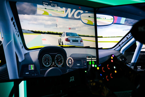 Blick aus einem Simulator-Cockpit mit Tachoanzeige und Drehzahlenmesser. Durch die Windschutzscheibe sieht man eine Rennstrecke und einen weißen Mercedes, der überholt wird. 