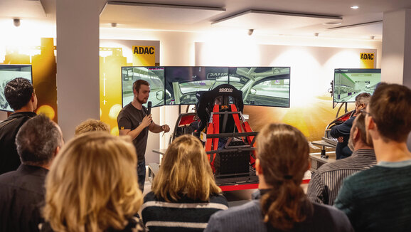 Mehrere Menschen die um unseren Leiter des SimRacing-Centers stehen und gespannt zuhören während er mit einem Mikrofon in der Hand eine Einweisung in unsere Full-Motion-Simulatoren gibt